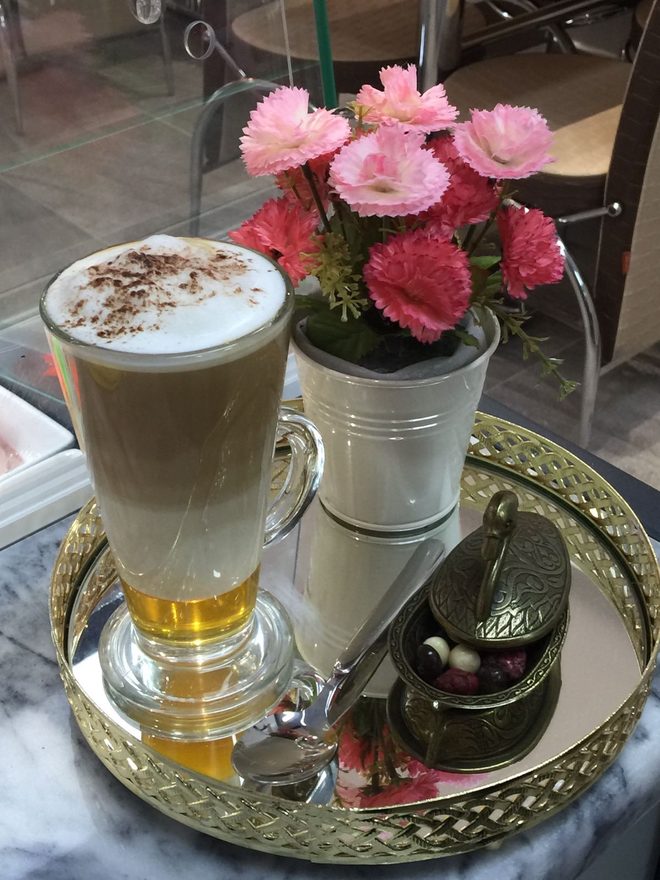 A tall glass latte on a brass tray beside a vase of pink carnations